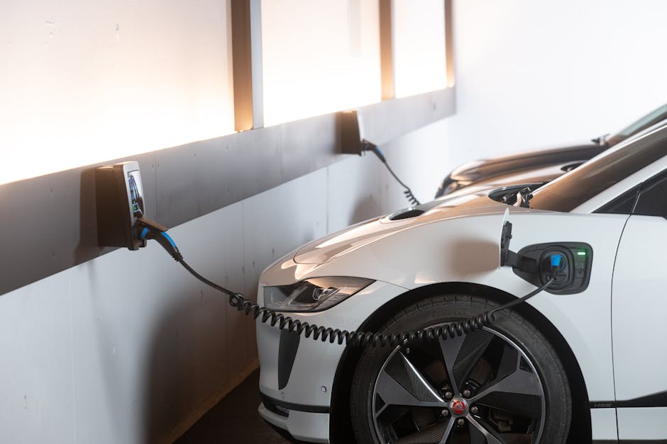 A white electric car is parked in a designated charging bay inside a garage or parking facility, connected to a wall-mounted charging station via a coiled black cable plugged into the vehicle's charging port on the front fender. The charging station features an electronic display and controls, positioned on a wall panel. The environment is well-lit with natural light coming through high windows, and the car appears to be in the process of charging. Visible surrounding objects include the vehicle's sleek, modern design and large alloy wheels, with additional similar vehicles parked nearby. This scene illustrates the typical setup for electric vehicle home or commercial charging, relevant to home relocation or vehicle transport services, such as those provided by Man and Van Hither Green, which handle comprehensive moving and transport logistics.