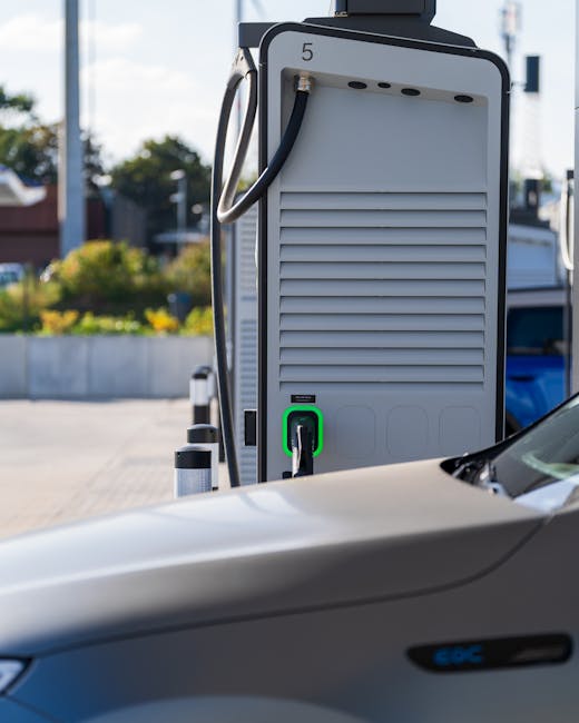 Close-up view of an electric vehicle charging station outdoors with a grey EV plugged into the socket, featuring a black cable secured to a side handle. The surrounding environment includes a paved area with bollards and black-and-white striped safety posts, and partially visible grassy areas with bushes and trees in the background. The charging station is numbered '5' at the top and has ventilation slats on the front. The scene appears to be part of a property or parking space suitable for home relocation or vehicle transportation purposes, with natural daylight illuminating the scene, consistent with professional moving or removal services provided by Man and Van Hither Green.