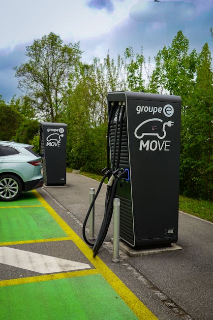 A white electric car is parked in a designated charging bay inside a garage or parking facility, connected to a wall-mounted charging station via a coiled black cable plugged into the vehicle's charging port on the front fender. The charging station features an electronic display and controls, positioned on a wall panel. The environment is well-lit with natural light coming through high windows, and the car appears to be in the process of charging. Visible surrounding objects include the vehicle's sleek, modern design and large alloy wheels, with additional similar vehicles parked nearby. This scene illustrates the typical setup for electric vehicle home or commercial charging, relevant to home relocation or vehicle transport services, such as those provided by Man and Van Hither Green, which handle comprehensive moving and transport logistics.