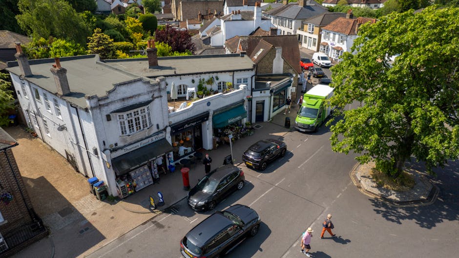 An aerial view of a small commercial building in Hither Green with a white facade and a teal awning, featuring a shopfront and flower displays outside. Several vehicles, including a black car, a green