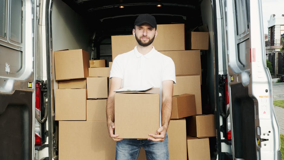 A man wearing a black cap, white polo shirt, and jeans stands inside the open rear of a large delivery van, holding a medium-sized cardboard box with both hands. The van's interior is filled with stacked cardboard boxes of various sizes, some wrapped in plastic or packing materials, positioned on the floor and along the sides. The setting appears to be outdoors, with a residential street visible in the background, including a sidewalk, trees, and neighboring houses. The lighting is natural, highlighting the details of the moving supplies and equipment, which include the boxes, packing materials, and the loading area of the van. This scene illustrates the process of home relocation and furniture transport, common in house removals and moving services offered by Man and Van Hither Green, emphasizing organized packing, loading, and transportation steps in a professional moving operation.