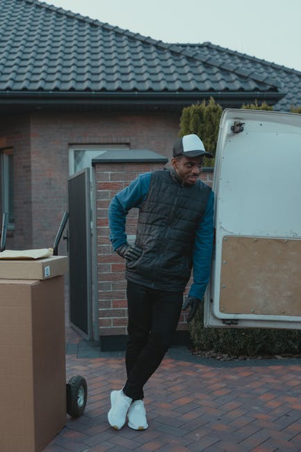 A man standing outside a residential property next to a white delivery van during daytime, wearing a baseball cap, a black padded vest over a blue long-sleeved shirt, black trousers, and white footwear. He is positioned on a paved driveway near a brick house with a grey tiled roof, with a partially open side door and a small green bush visible. To his left, there are stacked cardboard boxes and a wheeled trolley. The scene reflects the process of home relocation, with the man preparing to load or unload household items as part of a furniture transport or packing and moving operation managed by Man and Van Hither Green, a removal service focusing on local house moves and logistics.