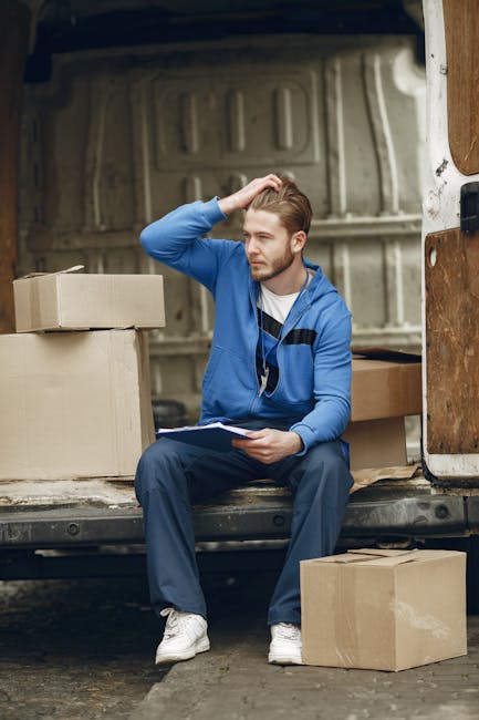 A man wearing a black cap, white polo shirt, and jeans stands inside the open rear of a large delivery van, holding a medium-sized cardboard box with both hands. The van's interior is filled with stacked cardboard boxes of various sizes, some wrapped in plastic or packing materials, positioned on the floor and along the sides. The setting appears to be outdoors, with a residential street visible in the background, including a sidewalk, trees, and neighboring houses. The lighting is natural, highlighting the details of the moving supplies and equipment, which include the boxes, packing materials, and the loading area of the van. This scene illustrates the process of home relocation and furniture transport, common in house removals and moving services offered by Man and Van Hither Green, emphasizing organized packing, loading, and transportation steps in a professional moving operation.