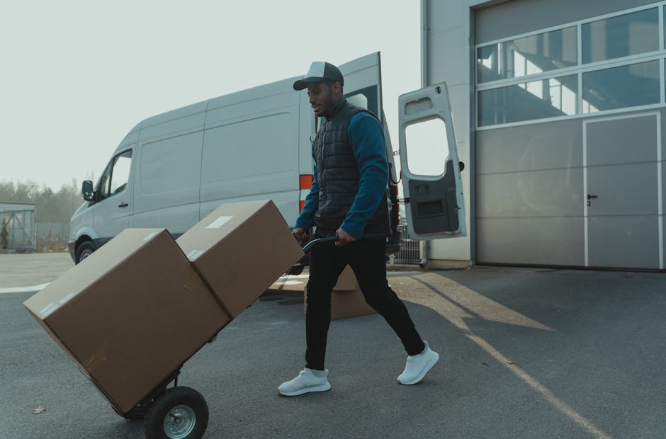 Two men are engaged in a home relocation process outdoors on a paved driveway, with one man lifting a cardboard box wrapped in plastic and labeled as fragile, while the other man steadies it. They are
