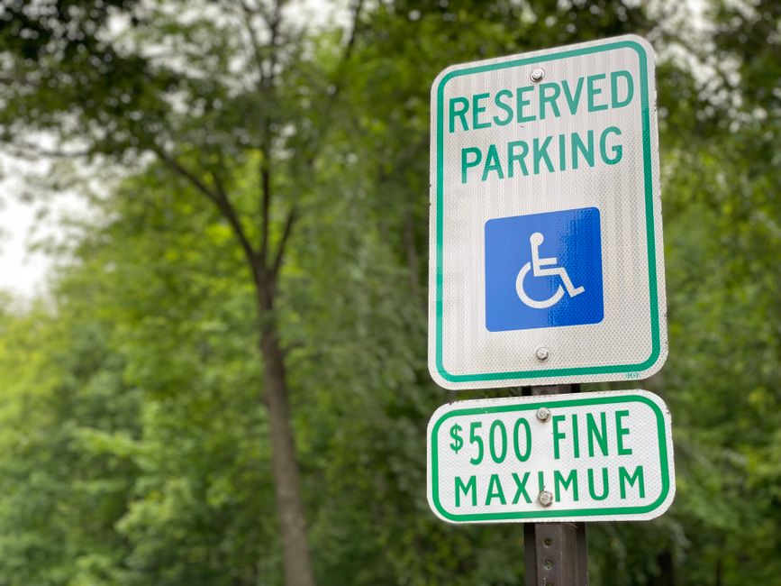 A close-up of a white reserved parking sign with green borders and text indicating parking for disabled individuals, featuring a blue wheelchair symbol. Below, an additional smaller sign specifies a maximum fine of $500 for violations. The signs are mounted on a metal post situated on a paved area adjacent to a property, with lush green trees and foliage in the background. The scene is set during daylight with natural lighting, and the signs are positioned in an outdoor environment consistent with urban or suburban parking regulations. As part of home relocation or furniture transport logistics, these signs are relevant for ensuring compliance with parking restrictions during removals, handled by Man and Van Hither Green.