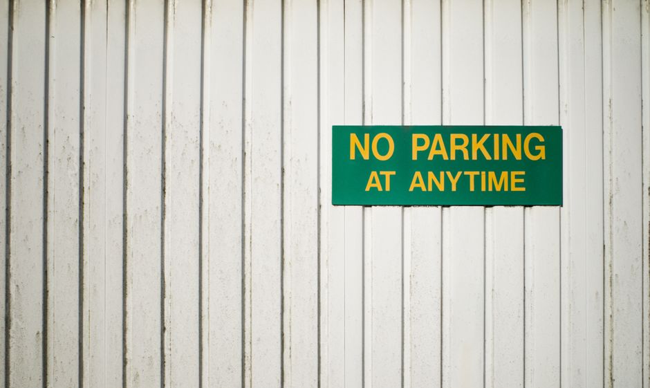 A close-up image of a white wooden wall made of vertical panels, displaying a green rectangular sign with bold yellow and white text that reads 'NO PARKING AT ANYTIME.' The sign is mounted securely on the wall, with no other objects or signage visible nearby. The lighting is natural and even, highlighting the texture of the wood and the contrasting colors of the sign. This image could be relevant for a house removal or moving service page discussing parking restrictions and compliance during home relocation in Hither Green, with [COMPANY_NAME] assisting in efficient furniture transport and packing logistics in accordance with local parking regulations.