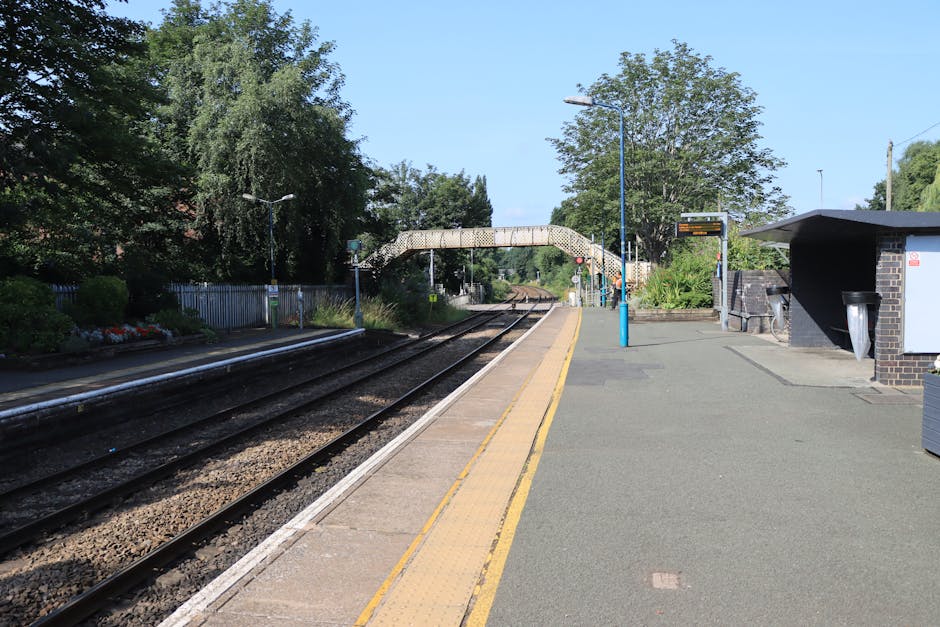 A quiet train station platform with two parallel railway tracks running alongside a paved pedestrian area. The platform edges are marked with a yellow tactile strip for visually impaired passengers, and the surface is a mix of concrete and asphalt. On the right side, there is a small brick building with a grey roof and a bin attached, and nearby a blue lamppost. A metal footbridge with a curved, perforated metal railing spans across the tracks in the background. The surrounding environment includes green trees and bushes, with signage indicating railway and station information visible near the footbridge. The scene is illuminated by natural sunlight, and there are no people visible on the platform or in the vicinity. This setting reflects typical elements of a local railway station, suitable for illustrating house removals involving transport to or from a train depot or station platform, as managed by Man and Van Hither Green.