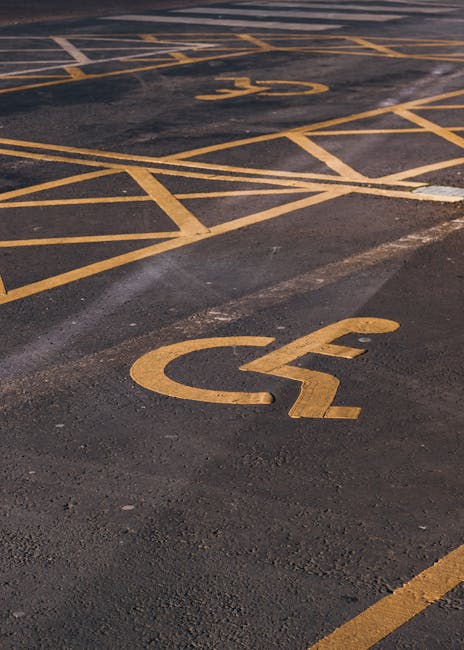 A close-up view of a parking lot surface with multiple yellow-painted parking space markings, including two visible disabled parking bays with the international wheelchair symbol. The parking area is made of dark asphalt with some signs of wear and patching. In the background, a portion of a white van is partially visible, indicating a vehicle used for home relocation and furniture transport. This setting, captured for the Hither Green area, illustrates the type of parking environment where Man and Van Hither Green’s removal services, as referenced in the Hither Green Village access guide, are commonly coordinated for efficient loading and unloading during house removals or moving projects. The lighting suggests daytime conditions with a natural, even light that highlights the clarity of the markings and surface details.