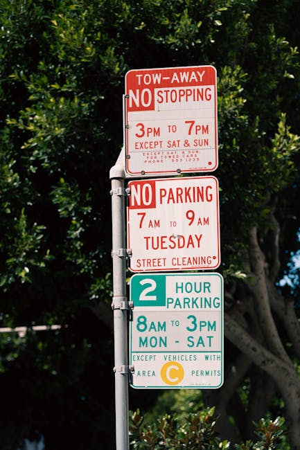 A vertical street signpost with four distinct signs affixed to a metal pole, located outdoors against a background of green foliage and a partly cloudy sky. The top sign is red and white, indicating a tow-away zone with no stopping permitted from 3pm to 7pm, except on Saturdays and Sundays. Below it, another red and white sign restricts parking from 7am to 9am on Tuesdays for street cleaning. The third sign is teal and white, displaying a two-hour parking limit from 8am to 3pm, Monday through Saturday, with an exception for vehicles with area C permits. The bottom sign, also teal and white, indicates additional parking regulations for a specific zone, again from 8am to 3pm, Monday through Saturday. The signs are clearly visible, with sunlight illuminating the signs and the natural environment surrounding the pole, used during home relocation or furniture transport planning to understand parking restrictions associated with Lewisham Council permit rules for Hither Green removals.