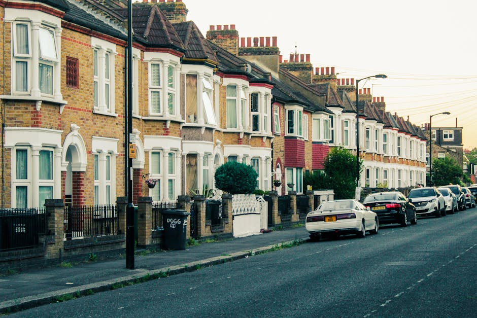 A row of traditional terraced houses in Hither Green with brick facades, bay windows, and small front gardens enclosed by low brick walls and black metal fences, situated alongside a street with parked cars on the pavement. Visible in the image are several cardboard boxes and wrapped furniture placed near the houses, indicating a home relocation process involving packing materials and furniture transport. The scene includes a man and van service van, positioned on the street close to the properties, demonstrating the loading or unloading phase of a removal. The background shows a clear sky with soft lighting, typical of early evening, and the street is equipped with streetlights and utility poles, with some greenery from small trees in front of the houses, aligning with local removal and moving logistics.