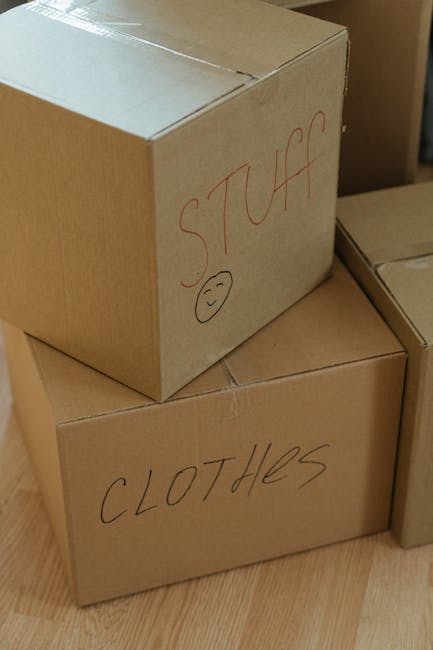 Close-up image of three cardboard moving boxes stacked inside a home, with the top box labeled 'STUFF' in red marker and a smiley face drawn underneath, while the lower box is labeled 'CLOTHES'. The boxes are positioned on a wooden floor, indicating a house relocation process. The boxes are sealed with packing tape, and the environment suggests an interior room during packing or unpacking for a house move. The labeling on the boxes reflects typical organisation in preparation for a moving and relocation service. This scene is representative of the packing stage often managed by companies like Man and Van Hither Green during home removals, focusing on packing and furniture transport logistics.