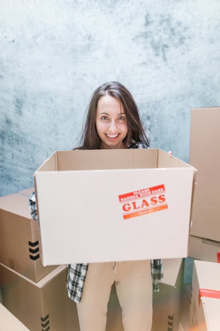 A young woman with long brown hair smiling at the camera while holding an empty cardboard box labeled 'Glass' in a room with light blue textured walls. She is wearing a casual plaid shirt and beige trousers. Around her, there are several other cardboard boxes stacked on the floor, some with printed arrow symbols and tape, indicating packing for a home relocation. The scene suggests the process of packing belongings in preparation for a house move, with the boxes placed in an organized manner inside a residential space. This image exemplifies the packing and moving stage of a house removal service, with the woman representing professional movers or a homeowner handling the packing process. The environment appears well-lit, emphasizing the organized nature of the packing activity.
