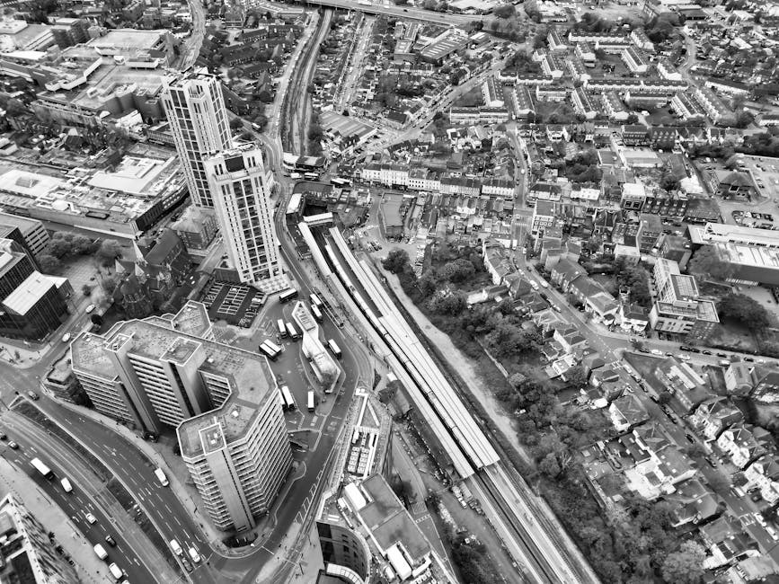Black and white aerial photograph of a densely built urban area showing high-rise office buildings, smaller commercial and residential structures, and streets with visible traffic. A railway station with platforms and train tracks runs through the scene, with platforms visible alongside the tracks. Adjacent roads are busy with cars and buses, with some vehicles waiting at traffic signals. The environment includes trees, parking areas, and open spaces interspersed among the buildings. The photograph captures the cityscape during daytime with natural lighting, highlighting the architectural layout and transportation infrastructure. This image background suggests moving logistics, such as home relocation or furniture transport, reflecting the busy urban setting that [COMPANY_NAME] might operate within as part of their removals or moving services.