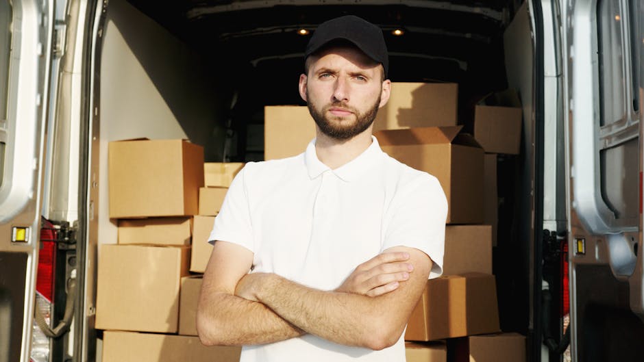 A man with a neutral expression, wearing a black cap and a white polo shirt, stands inside the open rear of a large van loaded with multiple cardboard boxes of various sizes. The boxes are arranged in the back of the vehicle, some stacked on top of each other, and are secured for a home relocation process. The van's interior is visible, with the sides and flooring made of metallic material, and the background showing more boxes towards the front. The vehicle is parked on a paved area, possibly outside a residence or commercial property, during daylight. This scene illustrates a furniture transport or packing and moving operation, with Man and Van Hither Green providing removals services, including efficient loading and transportation for same-day move requirements in Hither Green.