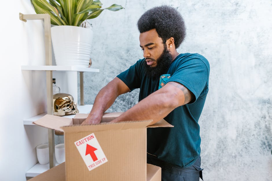 A man with a dark curly afro hairstyle and a beard is standing inside a home, opening a large cardboard moving box labeled with a red arrow and the words 'CAUTION THIS SIDE UP.' He is wearing a dark blue T-shirt and appears focused on packing or unpacking items during a house removal. The room features a white shelving unit mounted on a textured grey wall, with a large potted plant in a white ceramic planter on top, alongside a decorative brass skull ornament. Inside the box, visible packing materials include tissue paper and possibly fragile items, suggesting careful handling for a home relocation. The scene is well-lit with natural light, emphasizing the interior space for furniture transport and packing activities involved in the moving process. This image exemplifies the preparation and handling stages typical of professional removals, such as those provided by Man and Van Hither Green.