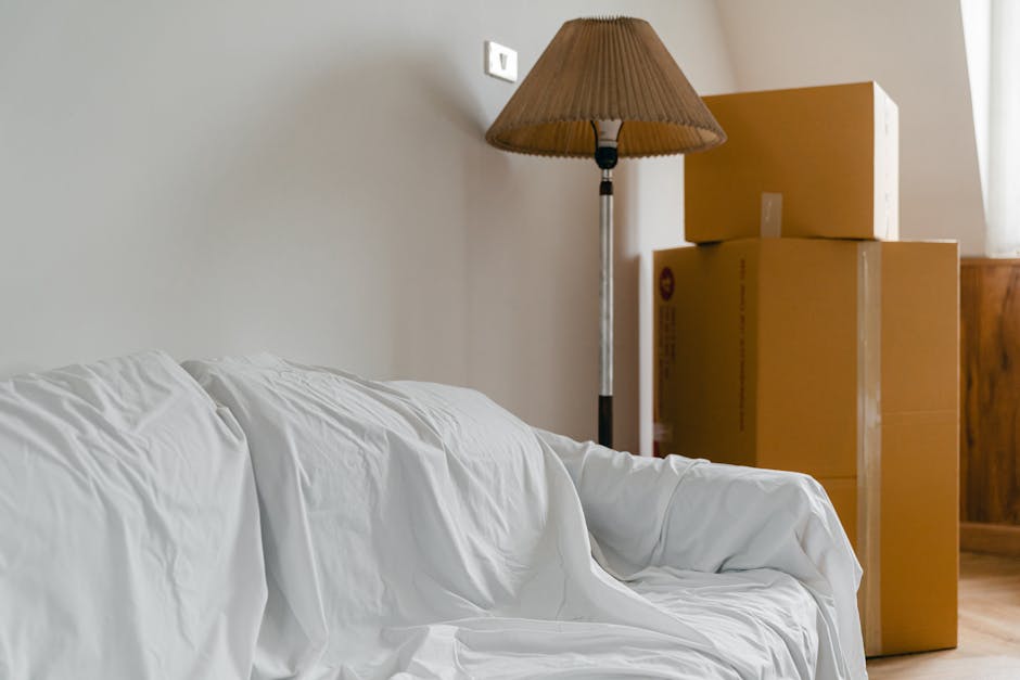 Inside a room prepared for a home relocation, cardboard boxes of various sizes stacked next to a wooden cabinet near a window, with natural light illuminating the space. A beige fabric-covered sofa with some creases is positioned in the foreground, partially blocking the view of the boxes. A tall, brown pleated lampshade on a metal stand is placed behind the boxes, adding ambient lighting. The image appears to be captured during the packing or loading process, with the boxes ready to be transported as part of furniture transport or moving activities. Man and Van Hither Green’s removal service may involve careful packing and loading in such a setting, typical for local house moves in SE13 or surrounding areas.