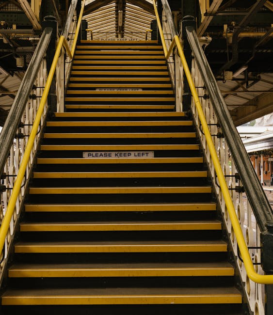 A set of industrial stairs with yellow handrails on either side, leading upwards in an indoor environment. The stairs are made of black treads with yellow edges and are marked with a white sign in the