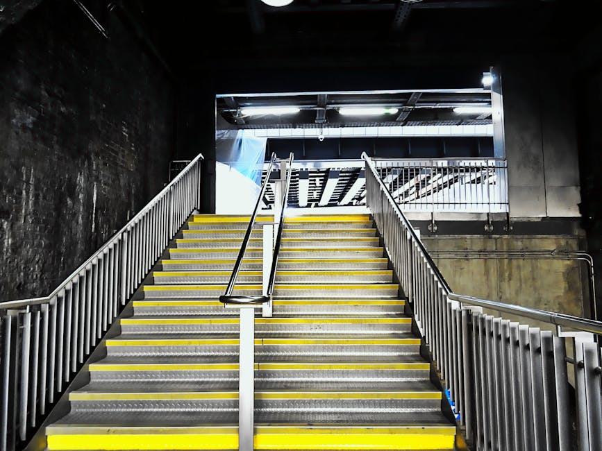A set of industrial stairs with yellow handrails on either side, leading upwards in an indoor environment. The stairs are made of black treads with yellow edges and are marked with a white sign in the