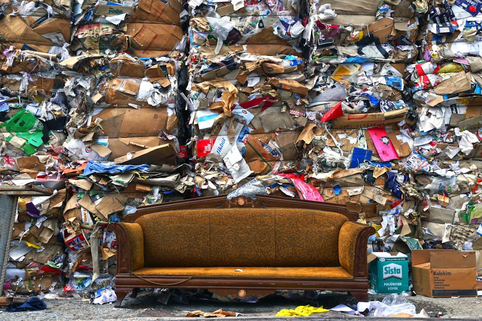 A vintage brown upholstered sofa with wooden armrests and decorative carvings is positioned on the pavement in front of a large, densely stacked pile of mixed household waste, including crumpled cardboard boxes, paper packaging, plastic wrappers, and various packaging materials. The background shows industrial waste collection or recycling site with the trash arranged in large bundles. The sofa is part of an initial home relocation or furniture transport process, with visible signs of wear and tear, and is situated near the entrance of a property or waste disposal area. The scene is well-lit, highlighting the contrast between the furniture and the cluttered backdrop, which encapsulates the challenges of bulky object disposal in Lewisham, processed by a removal company like Man and Van Hither Green, specializing in removals and waste clearance.