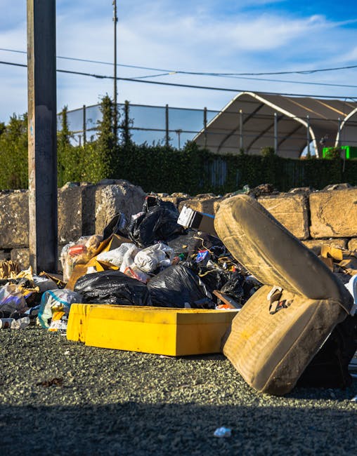 A mound of mixed household waste and household items, including black and white plastic garbage bags, a yellow plastic storage box, a large used beige fabric-upholstered armchair with visible stains and damage, and various discarded packaging materials, all situated on a gravel surface beside a stone wall and at the edge of a pavement. In the background, there are overhead power lines, a fence, trees, and a clear blue sky, indicating an outdoor setting. This scene illustrates waste disposal during a home relocation or furniture removal, with the items likely cleared from inside a property before transport or disposal, a common aspect of professional removals services offered by Man and Van Hither Green.