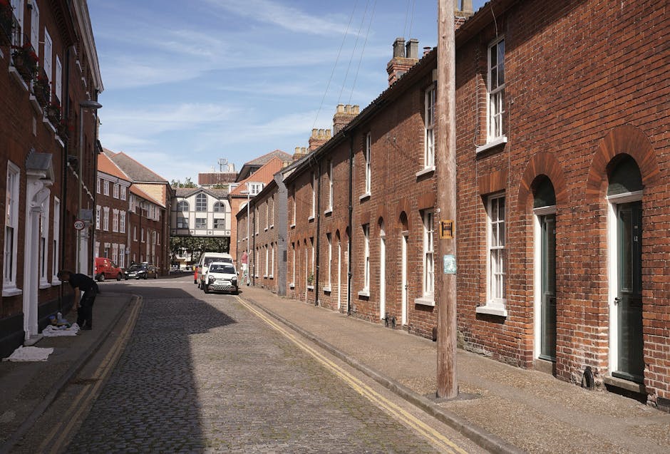 A narrow residential street lined with red-brick terraced houses, each with white-framed sash windows and small front doors. The cobblestone pavement is partially shaded by a wooden utility pole situated on the sidewalk, with overhead power lines running along the street. One individual is visible on the left, dressed in dark clothing, handling packaging or moving materials on the pavement, indicating packing or unpacking activity during a house relocation. A white van is parked further down the street, with the rear doors possibly open for loading or unloading furniture and boxes. The scene illustrates a typical urban setting suitable for a house removal service, with a clear blue sky overhead suggesting daytime conditions. In the background, a modern building with large windows spans the width of the street, reinforcing a mixed architectural environment. The image emphasizes tight street access considerations encountered by local moving companies such as Man and Van Hither Green during furniture transport and home relocation projects. The overall environment reflects the typical logistical challenges of executing efficient removals in narrow street settings.