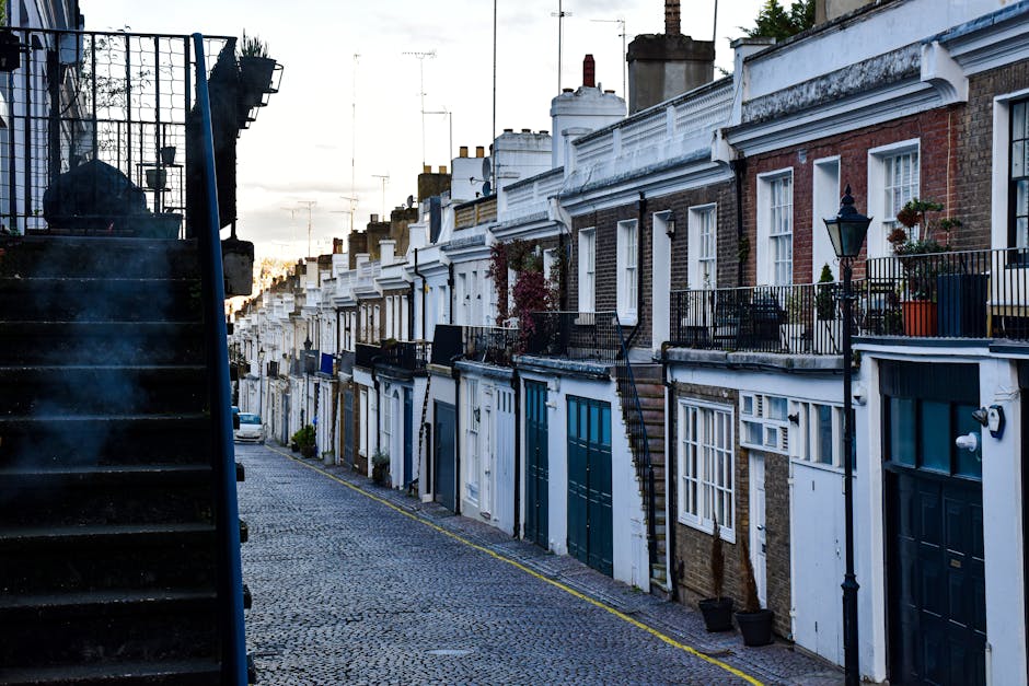 A narrow residential street lined with red-brick terraced houses, each with white-framed sash windows and small front doors. The cobblestone pavement is partially shaded by a wooden utility pole situated on the sidewalk, with overhead power lines running along the street. One individual is visible on the left, dressed in dark clothing, handling packaging or moving materials on the pavement, indicating packing or unpacking activity during a house relocation. A white van is parked further down the street, with the rear doors possibly open for loading or unloading furniture and boxes. The scene illustrates a typical urban setting suitable for a house removal service, with a clear blue sky overhead suggesting daytime conditions. In the background, a modern building with large windows spans the width of the street, reinforcing a mixed architectural environment. The image emphasizes tight street access considerations encountered by local moving companies such as Man and Van Hither Green during furniture transport and home relocation projects. The overall environment reflects the typical logistical challenges of executing efficient removals in narrow street settings.